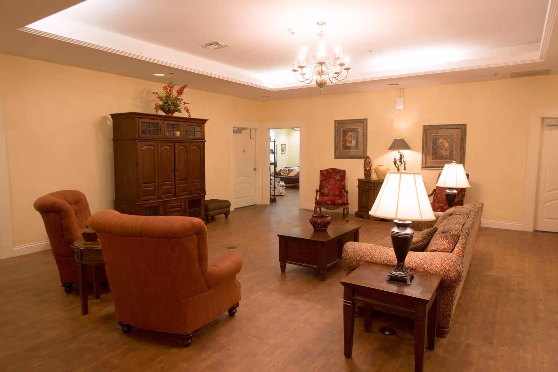 A spacious living room area with wooden flooring, beige walls, and a white ceiling with recessed lighting and a chandelier. The room is furnished with a patterned sofa, two red armchairs, a wooden coffee table, side tables with lamps, a wooden cabinet with a floral arrangement on top, and framed artwork on the walls. There are two doorways visible, one leading to another room with a bed.