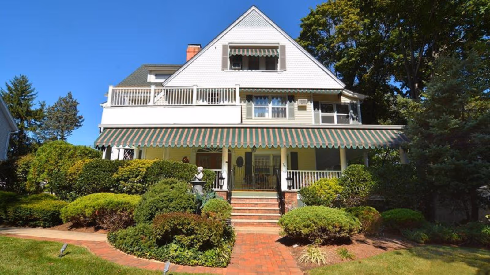 Front exterior view of a large, white, multi-story house with a green and red striped awning over the porch. The house is surrounded by well-maintained bushes and greenery under a clear blue sky.