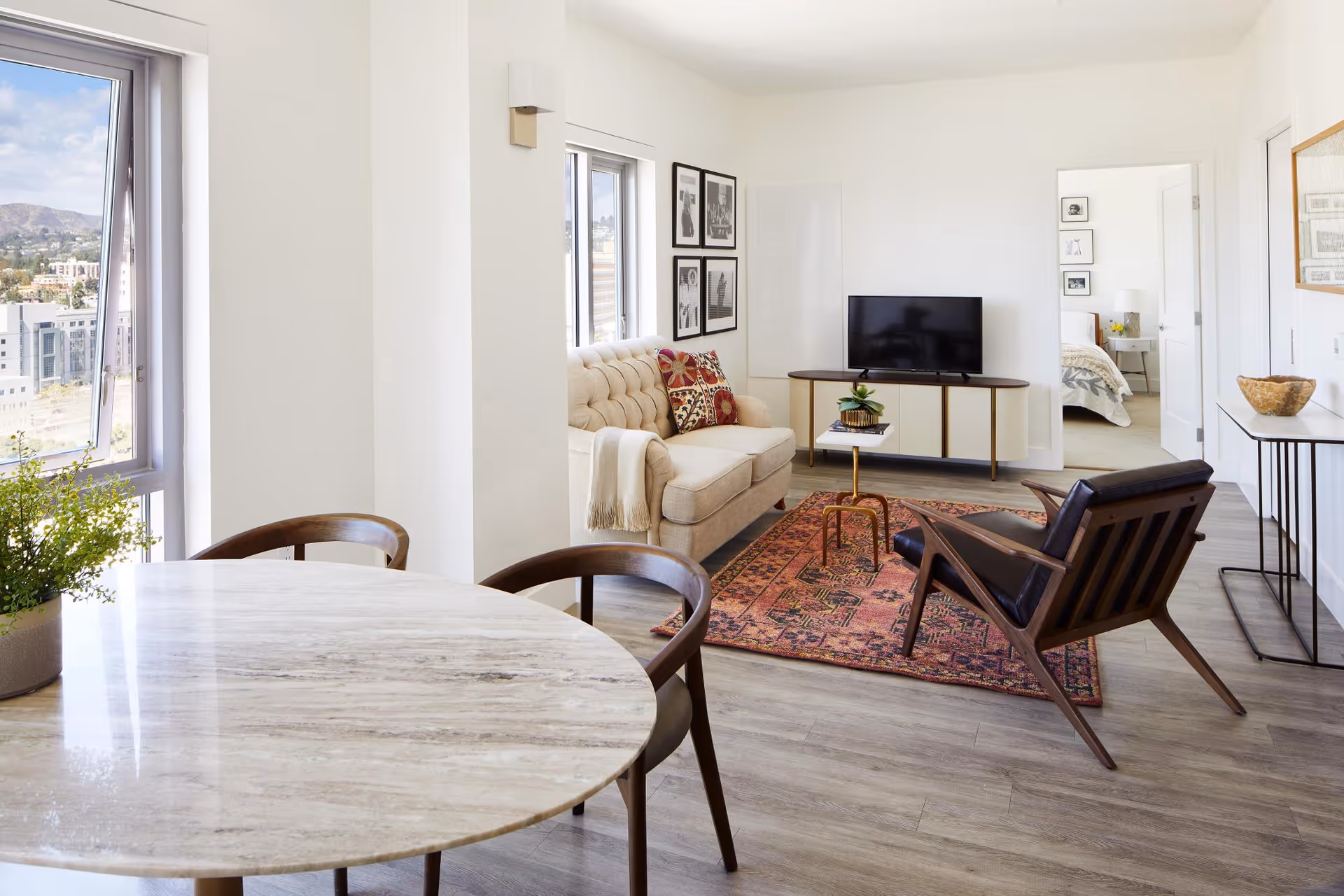 Bright open living room and dining area with a round marble table, beige sofa, patterned rug, wooden armchair, TV console, and a view into a bedroom.