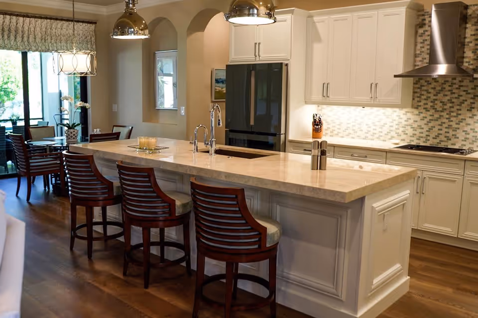 Modern kitchen with a large marble island countertop featuring three wooden bar stools with cushioned seats. The kitchen has white cabinetry, a stainless steel refrigerator, a built-in stovetop with a stainless steel range hood, and a tiled backsplash. In the background, there is a dining area with a round table and chairs, a large window with patterned curtains, and pendant lights hanging above the island and dining table.