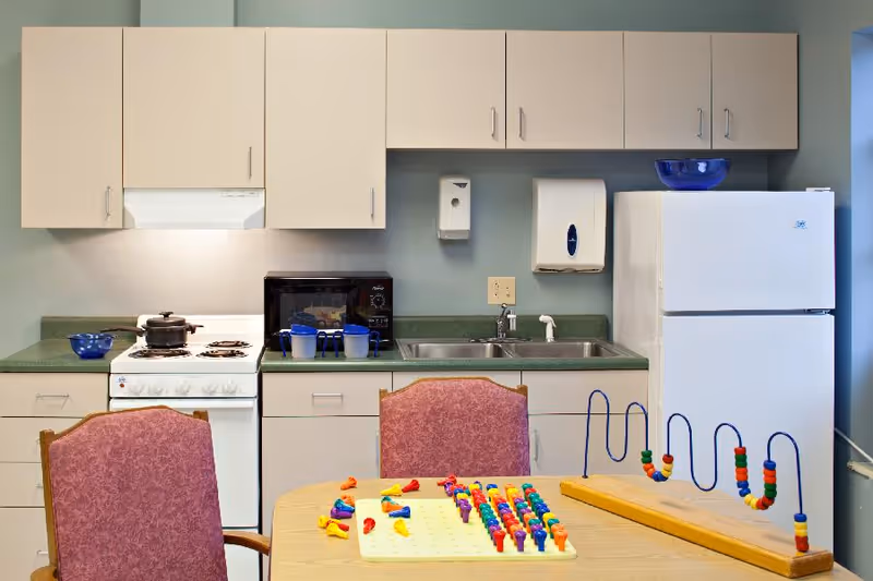 A small kitchen area with beige cabinets, a white stove with a pot on it, a microwave, a double sink, and a white refrigerator. In front of the kitchen is a table with two pink upholstered chairs and colorful children's toys on the table.