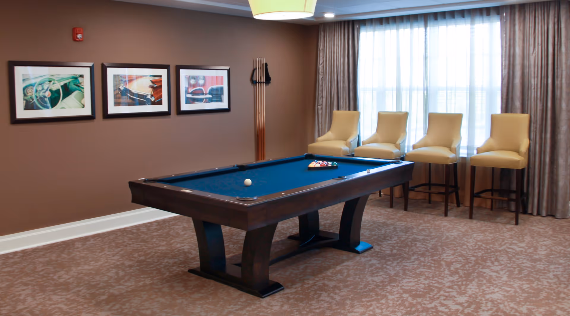 A room with a pool table in the center, four beige high-back chairs lined up against a window with curtains, and three framed pictures on a brown wall. The floor is carpeted with a patterned design.