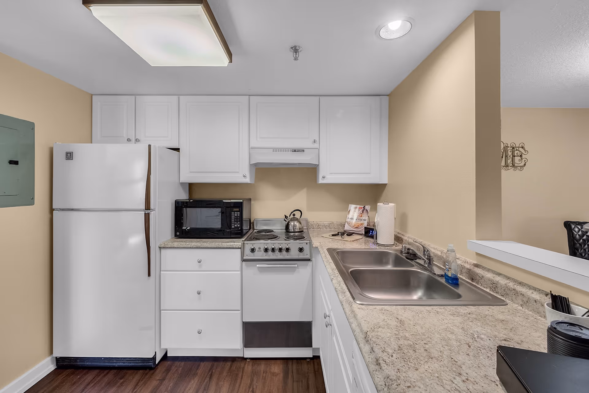 A compact kitchen with white cabinets, a white refrigerator, a black microwave, an electric stove with a kettle on top, a double stainless steel sink, and a beige countertop. The walls are painted beige and the floor has dark wood flooring.