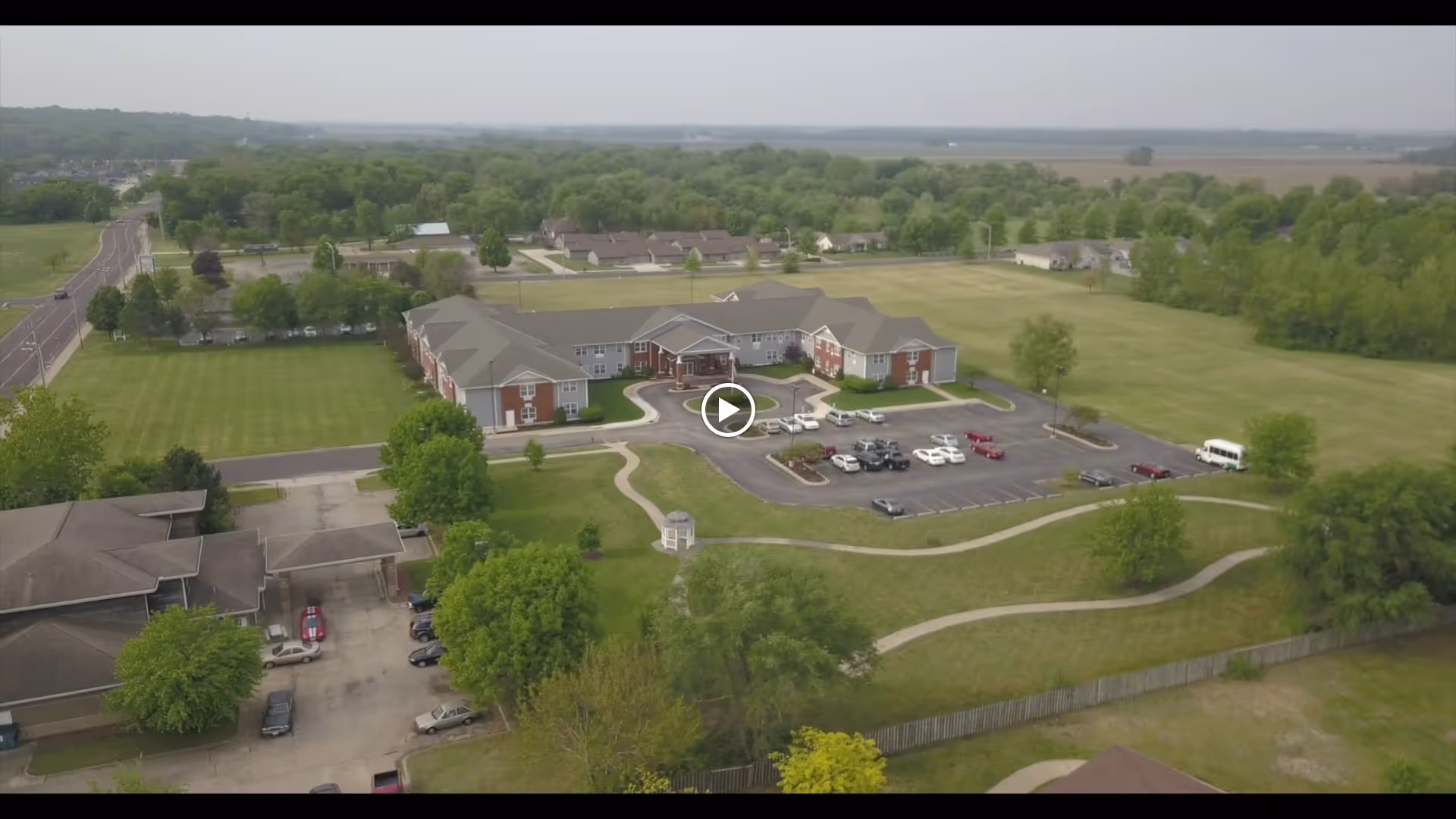 Aerial view of John M. Evans Supportive Living Community showing a large building with a parking lot, surrounded by green lawns, trees, and a winding pathway. The facility is located in a rural area with open fields and some residential houses in the distance.