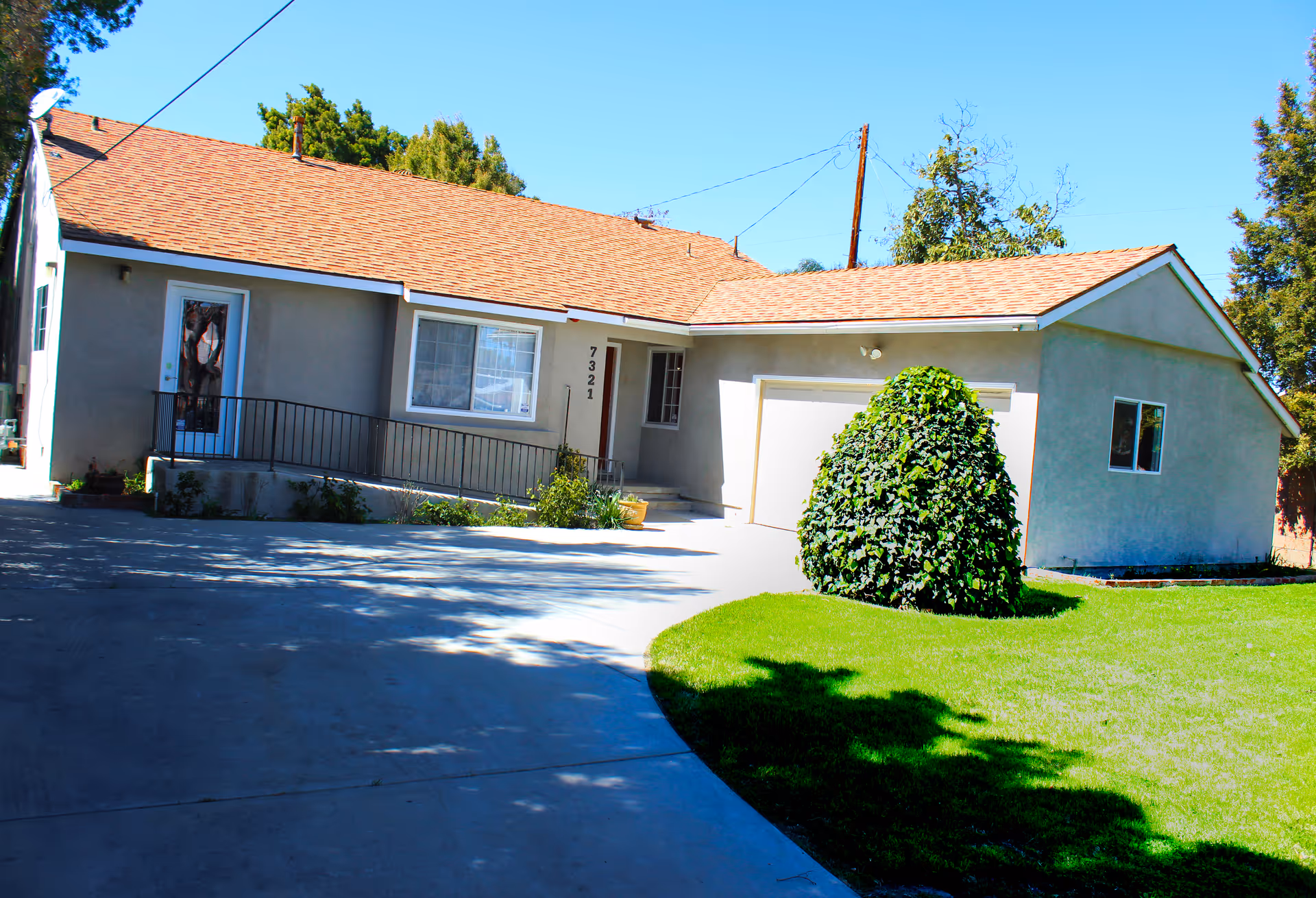 Exterior view of a single-story house with a beige exterior and a reddish-brown shingled roof. The house has a garage, a front door with a small ramp, a window, and a well-maintained lawn with a large bush. Trees and a clear blue sky are visible in the background.