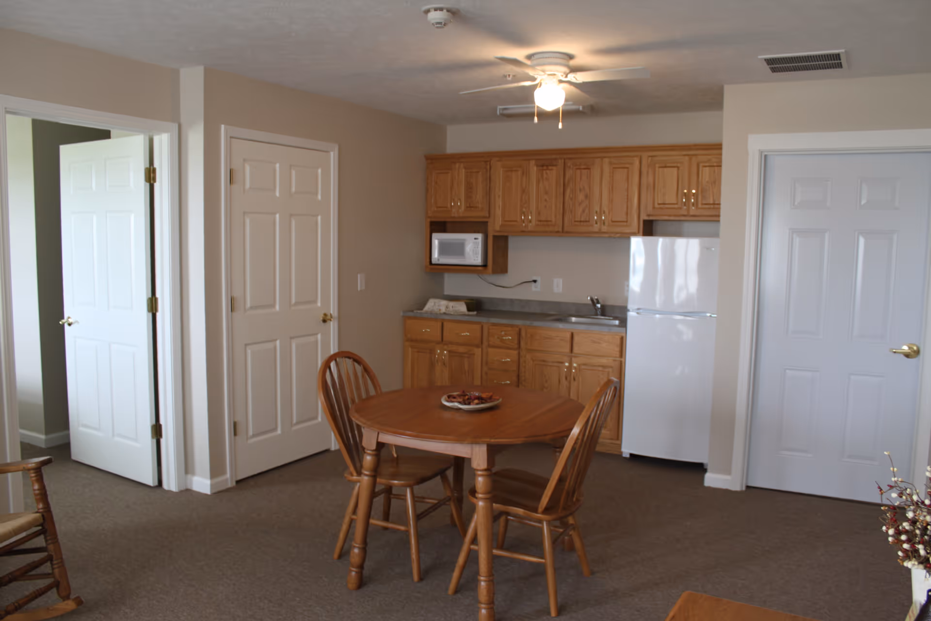 Interior view of a senior living facility kitchen and dining area with wooden cabinets, a white refrigerator, microwave, a round wooden table with two chairs, beige walls, and carpeted floor. There are three white doors visible and a ceiling fan with a light.