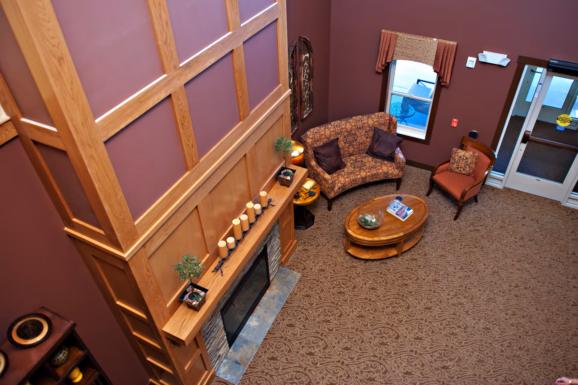 A cozy living room area with a patterned loveseat and armchair, a wooden coffee table with decorative items, a fireplace with candles and small plants on the mantel, and a window with a valance. The walls are painted a warm brown color and the carpet has an intricate design. The entrance door is visible in the background.