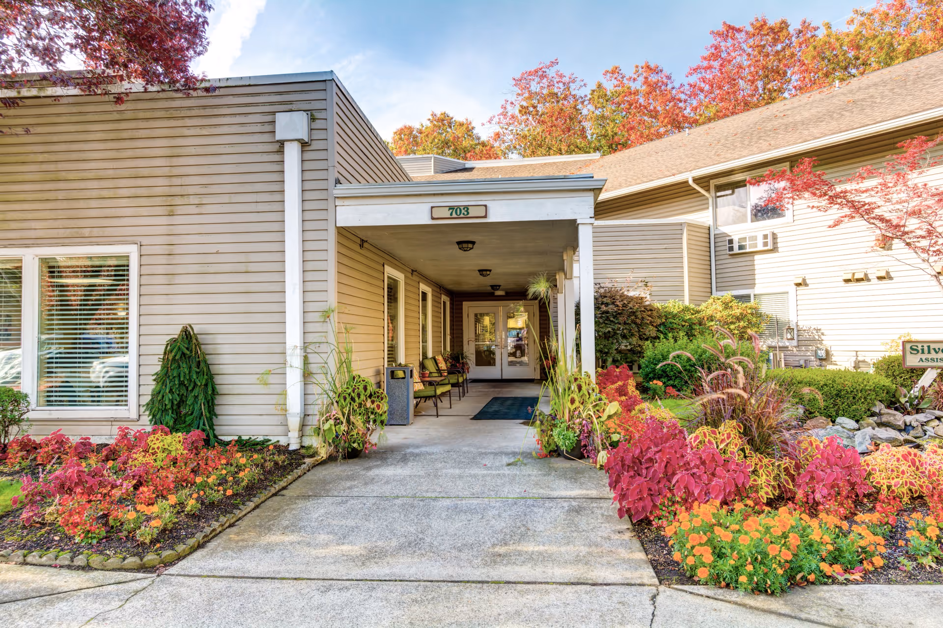 Covered main entrance of Silver Creek Senior Living with a walkway, seating under a porch, and vibrant flower beds.