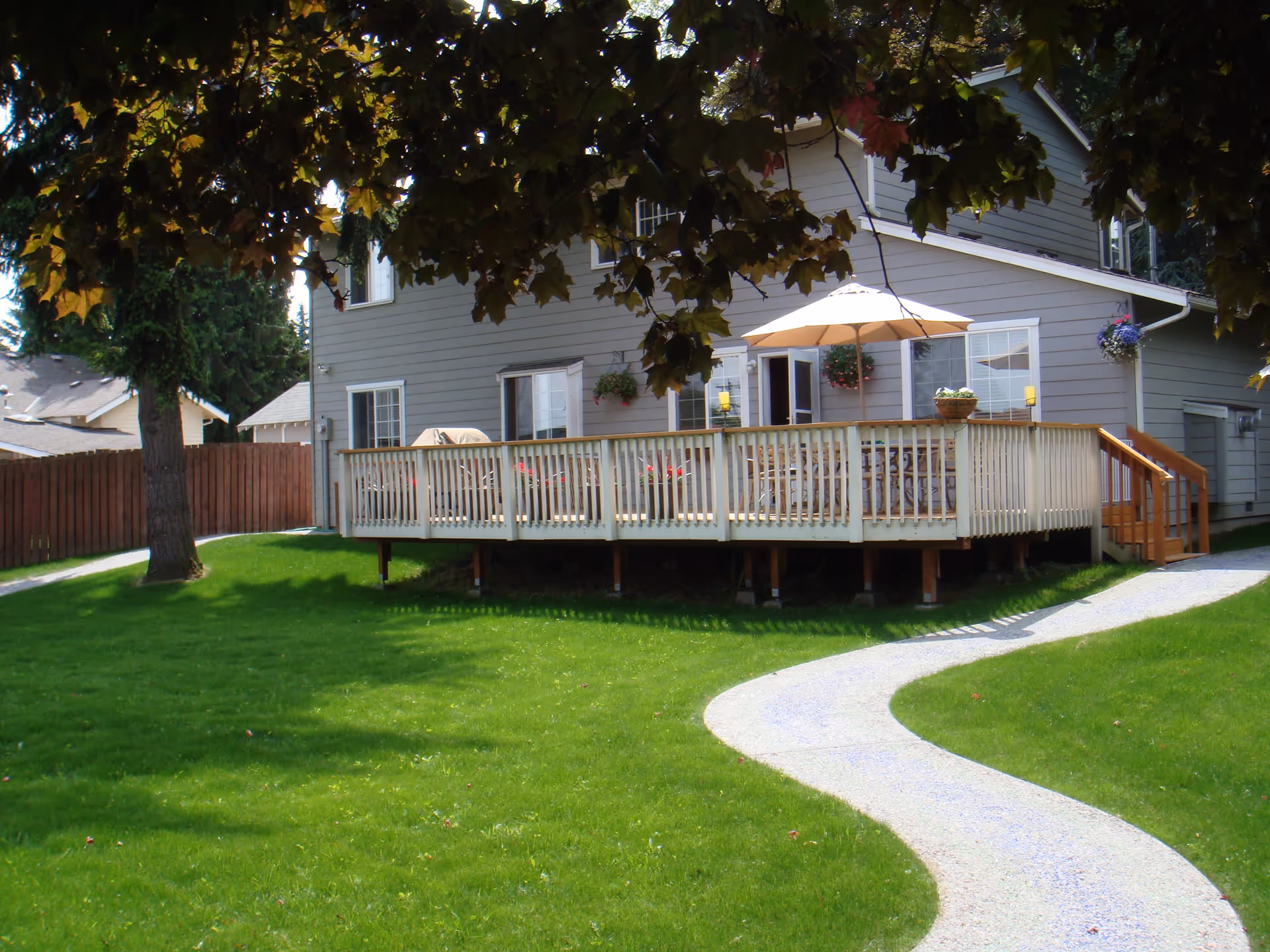 A gray two-story house with a raised wooden deck, patio umbrella, and a curved path across a green lawn.