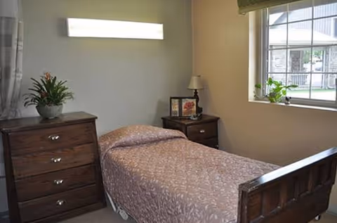 Bedroom in a senior living facility with a bed, two dressers, and a window.