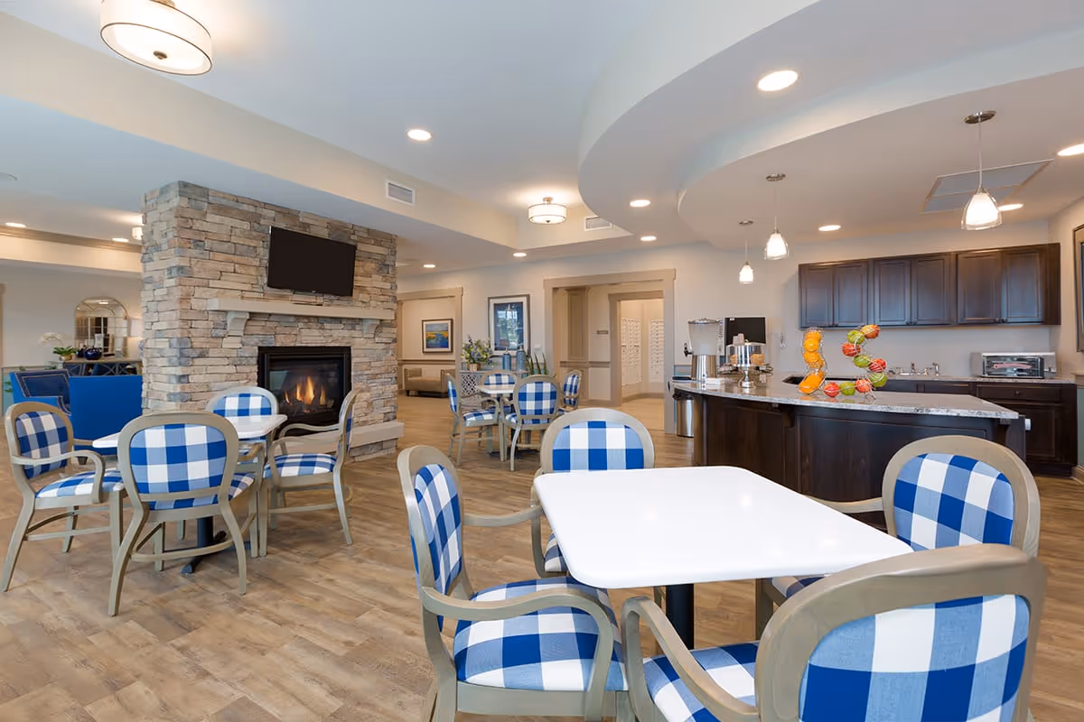 A bright and spacious common area in an assisted living facility featuring several tables with blue and white checkered chairs. There is a stone fireplace with a mounted TV above it, a kitchen area with dark wood cabinets and a countertop with fruit decorations, and modern lighting fixtures on the ceiling.