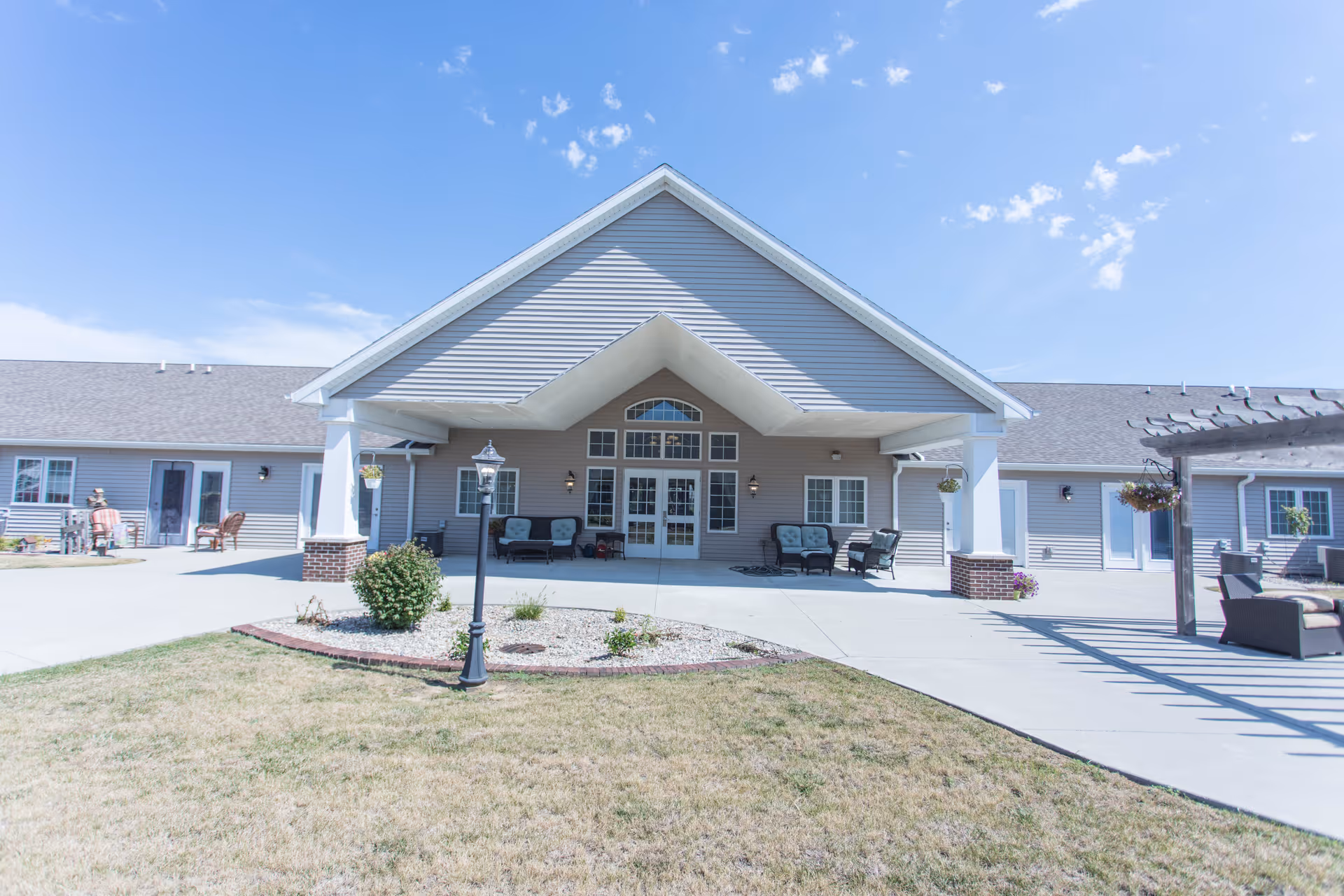 Exterior view of a single-story senior living facility building with a large covered entrance area. The building has gray siding, white trim, and multiple windows and doors. There are outdoor seating areas with chairs and small tables on the concrete patio. A small landscaped area with a lamp post and some plants is in front of the entrance. The sky is clear with a few clouds.