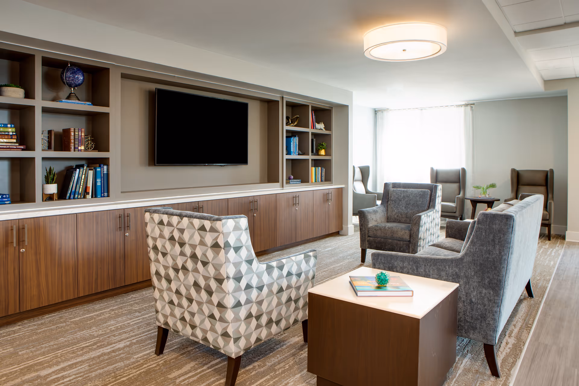 A modern living room area in a senior living facility with a patterned armchair, two gray armchairs, and a gray sofa arranged around a wooden coffee table with books and a decorative item on it. The room features built-in wooden shelves and cabinets with books and decorative items, a large flat-screen TV mounted on the wall, and large windows letting in natural light.