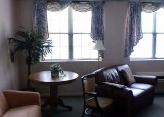 Small living room with a leather loveseat, upholstered chairs, a round wooden table with potted plants, a floor lamp, and large windows with floral curtains.
