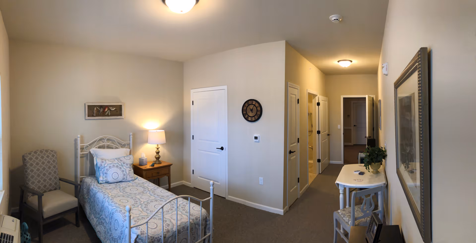 Small senior living bedroom with a single bed, bedside table and lamp, a chair, and a hallway leading to other rooms.