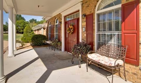 A covered outdoor porch area with brick walls, red front doors, and windows with red shutters. There are decorative plants in pots and cushioned metal benches along the porch. White columns support the roof, and greenery is visible in the background.