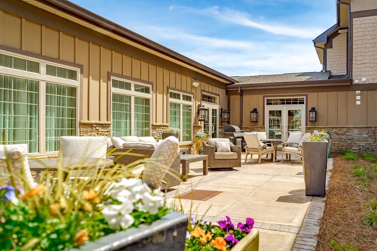Outdoor patio area at Celebration Village Forsyth featuring cushioned wicker chairs and sofas arranged around small tables, with large windows and doors on the building wall. Flower planters with colorful blooms are in the foreground, and the sky is clear and blue.