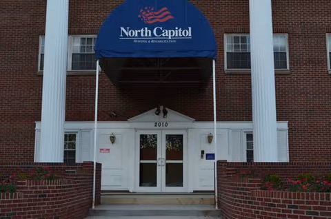 Front entrance of the North Capitol Nursing and Rehabilitation building with a blue awning, white double doors, and a brick facade.