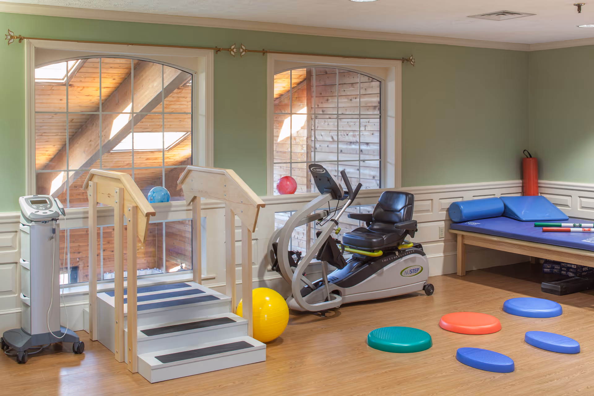 A senior living community exercise room with wooden flooring, two large windows showing a wooden exterior, a recumbent exercise bike, a small set of stairs with handrails, a yellow exercise ball, colorful balance pads on the floor, and a padded therapy table with blue cushions and exercise sticks.
