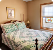Cozy bedroom with a wooden bed dressed in a green floral quilt, a nightstand and lamp, framed wall art, and a window with a decorative sun catcher.
