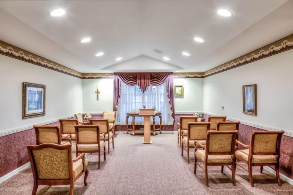 A small chapel or meditation room with rows of wooden chairs upholstered in gold fabric facing a wooden podium and a table with decorative items. The room has beige walls with a maroon and gold border, maroon curtains over a window, framed pictures on the walls, and a cross mounted on the left wall.