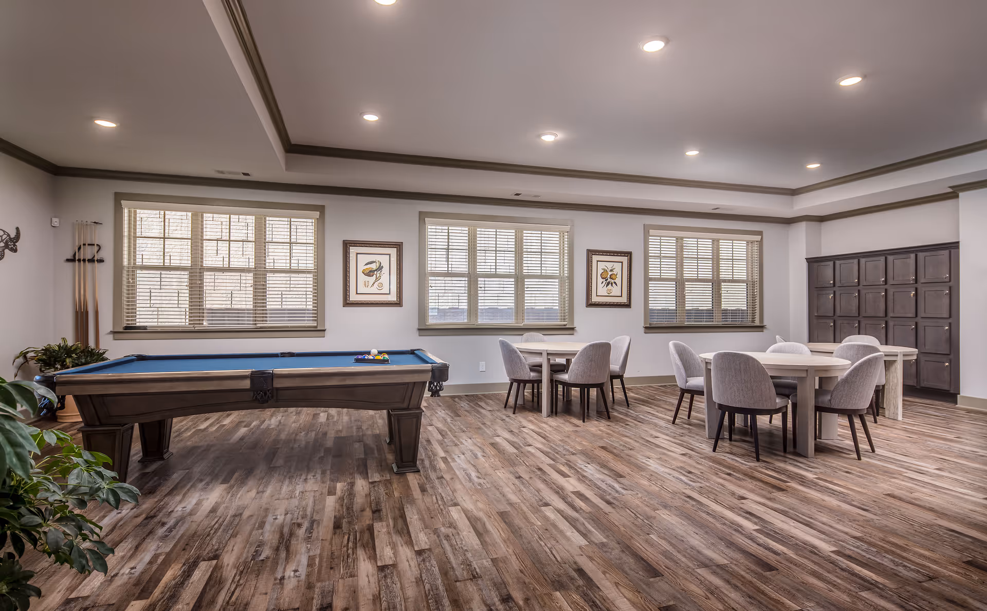 A spacious recreational room with a pool table on the left and two round tables with chairs on the right. The room has large windows with blinds, framed artwork on the walls, wood flooring, and a set of lockers in the back corner. Ceiling lights illuminate the room.
