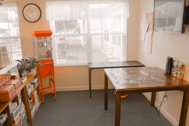 A cozy room with two tables, one holding an incomplete jigsaw puzzle. There is a bright orange popcorn machine on a stand near the window, a clock on the wall showing 11:55, a bookshelf filled with books and games, and a flat-screen TV mounted on the wall. The room has large windows with blinds letting in natural light.