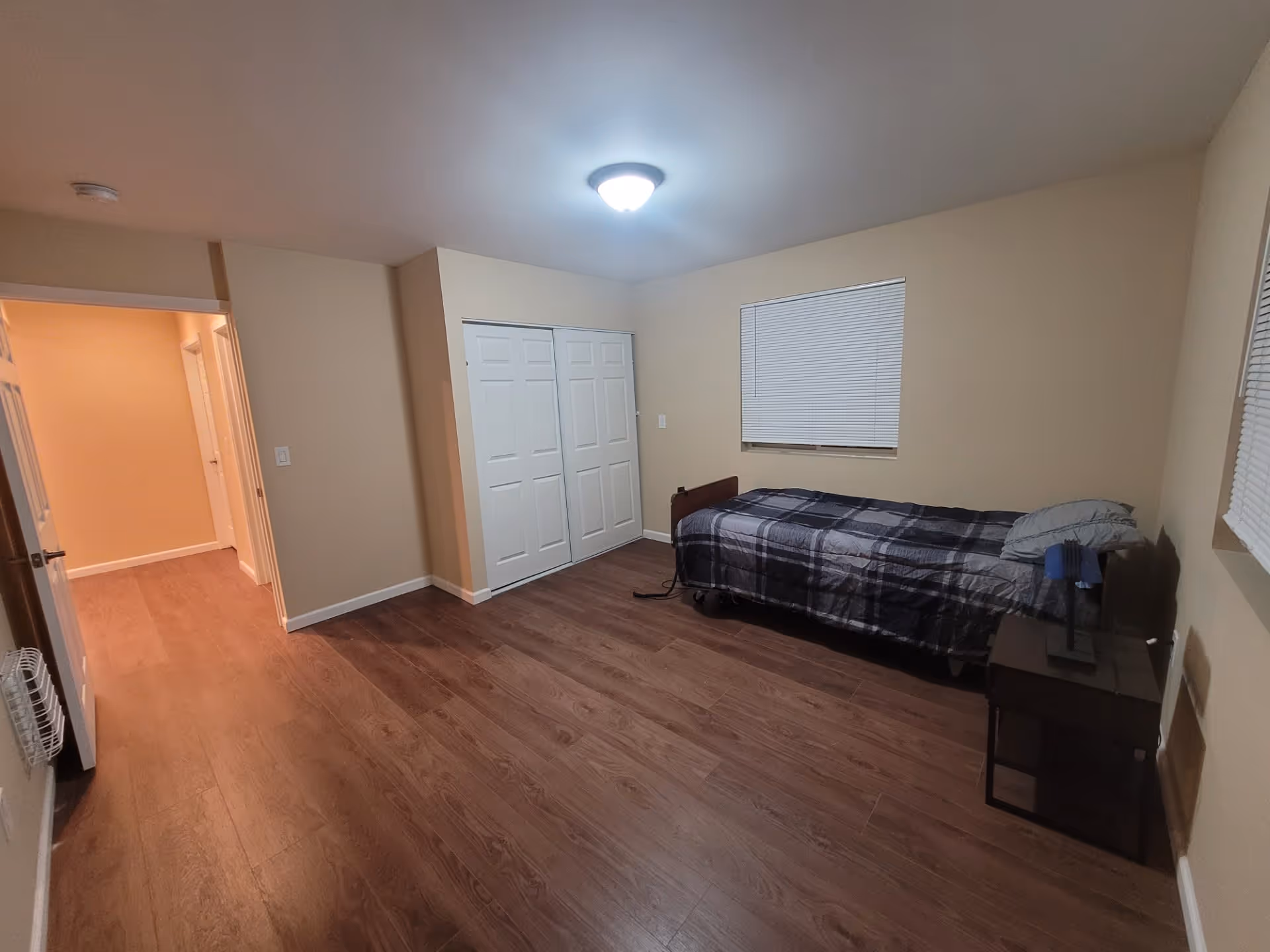 Simple bedroom with a single bed, nightstand, closed double-door closet, beige walls, and wood-look flooring under a ceiling light.