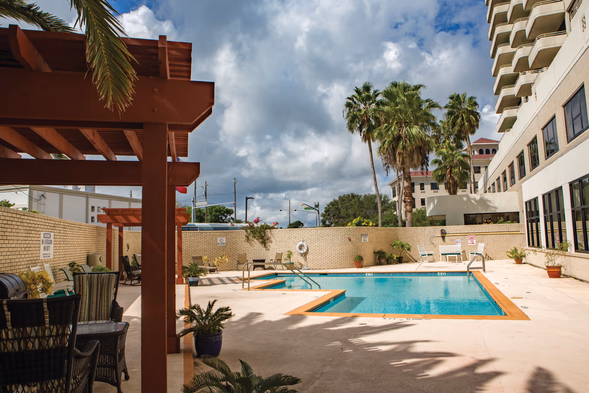 Outdoor swimming pool area with clear water, surrounded by a concrete deck and a beige brick wall. There are several potted plants and palm trees around the pool. On the left side, there is a shaded seating area with chairs and tables under wooden pergolas. A multi-story building with balconies is visible on the right side under a partly cloudy sky.