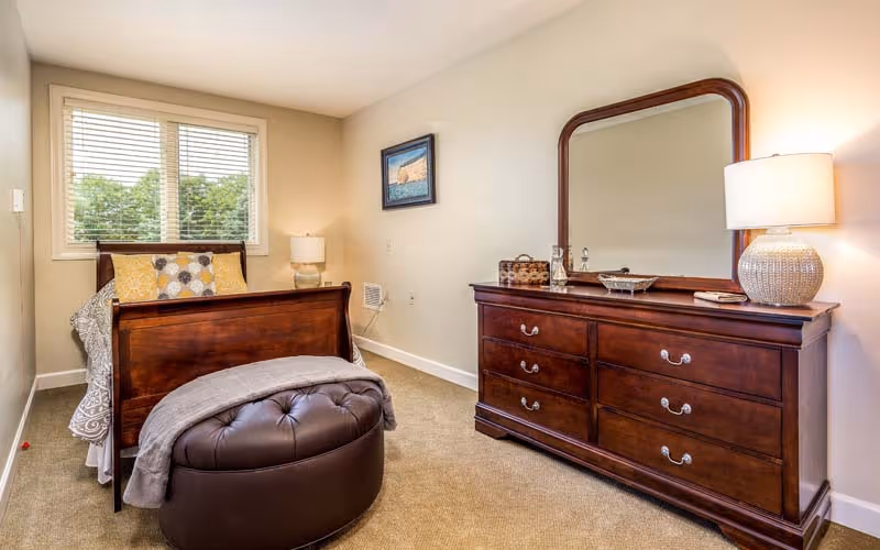 A cozy bedroom with a wooden sleigh bed, round leather ottoman, and a large wooden dresser with mirror and lamps.