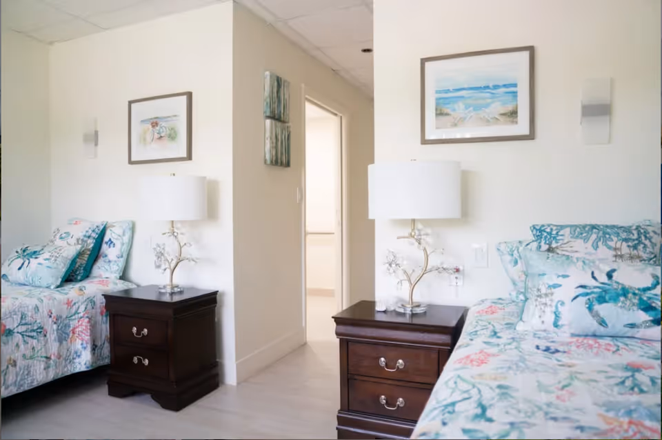 A bright bedroom with two twin beds separated by a dark wooden nightstand with a white lamp. The beds have floral and sea-themed bedding with multiple pillows. The walls are light-colored and decorated with framed beach-themed artwork. There is an open doorway leading to another room.