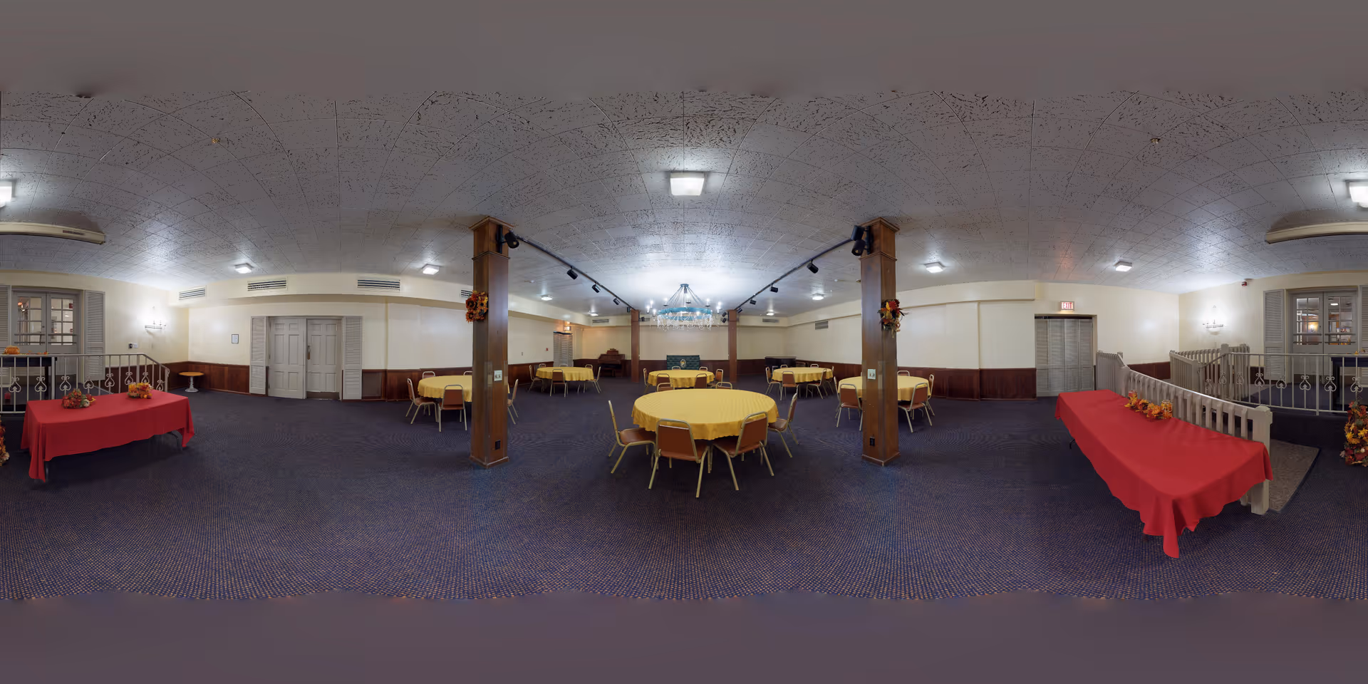 A large dining room with multiple round tables covered with yellow tablecloths and surrounded by chairs. Two rectangular tables with red tablecloths are placed against the walls, decorated with small autumn-themed centerpieces. The room has a blue carpet, white walls with wooden paneling on the lower half, and a white ceiling with multiple light fixtures. Two wooden pillars with fall decorations stand near the center of the room. There is a railing and a small staircase on the right side of the room.
