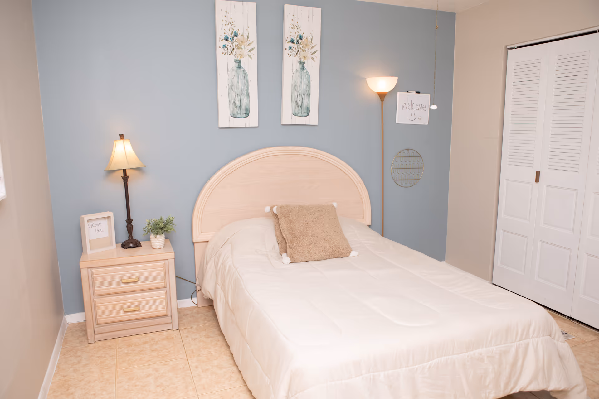 A cozy bedroom with a light wood bed frame and a beige comforter. There is a brown pillow on the bed. On the left side, there is a wooden nightstand with two drawers, a small lamp, a framed note that says 'Welcome Home', and a small potted plant. The wall behind the bed is painted blue and decorated with two vertical floral paintings. On the right side, there is a floor lamp with a whiteboard hanging from it that says 'Welcome' with a smiley face. A white closet door is visible on the right side of the room.