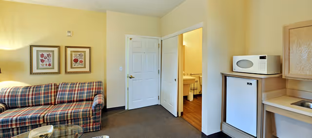 Small living area with a plaid sofa, a kitchenette with microwave and mini-fridge, and an open door revealing a bathroom.