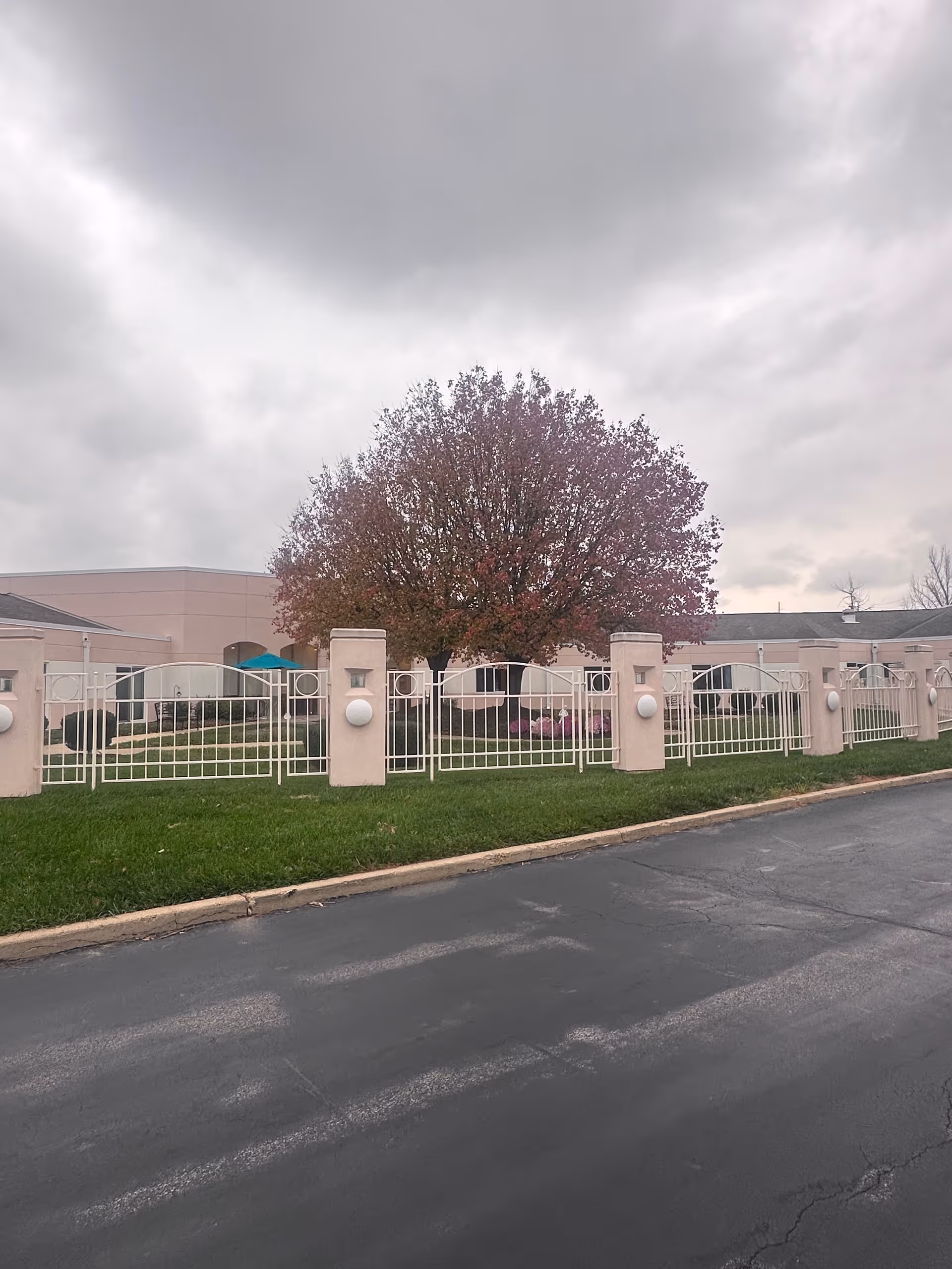Exterior view of a senior living facility named Garden Villas South, showing a pink building behind a white metal fence with pillars. There is a large tree with reddish leaves in the courtyard area, green grass, and a paved driveway in the foreground under a cloudy sky.