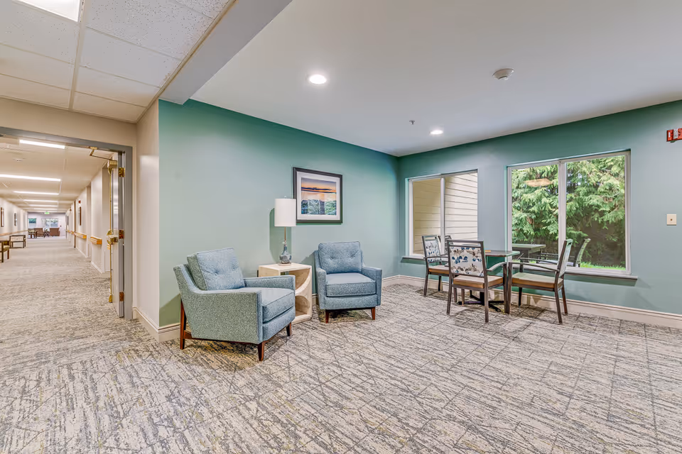 A cozy sitting area in a senior living facility with two blue upholstered armchairs, a small wooden side table with a lamp, and a framed picture on a green wall. Next to the sitting area is a dining table with four chairs near large windows showing greenery outside. A hallway extends to the left with more seating visible in the distance.
