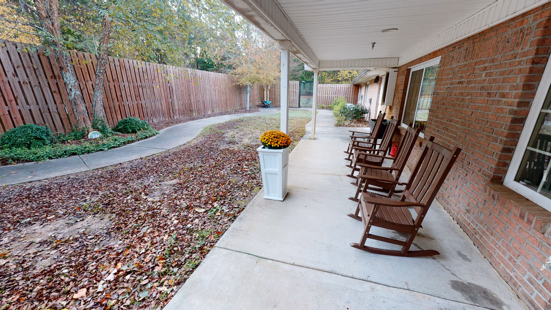 Covered outdoor patio area with several wooden rocking chairs lined up against a brick wall. A white planter with yellow flowers is placed on the concrete floor. The patio overlooks a fenced yard with a curved concrete pathway and some greenery along the fence.