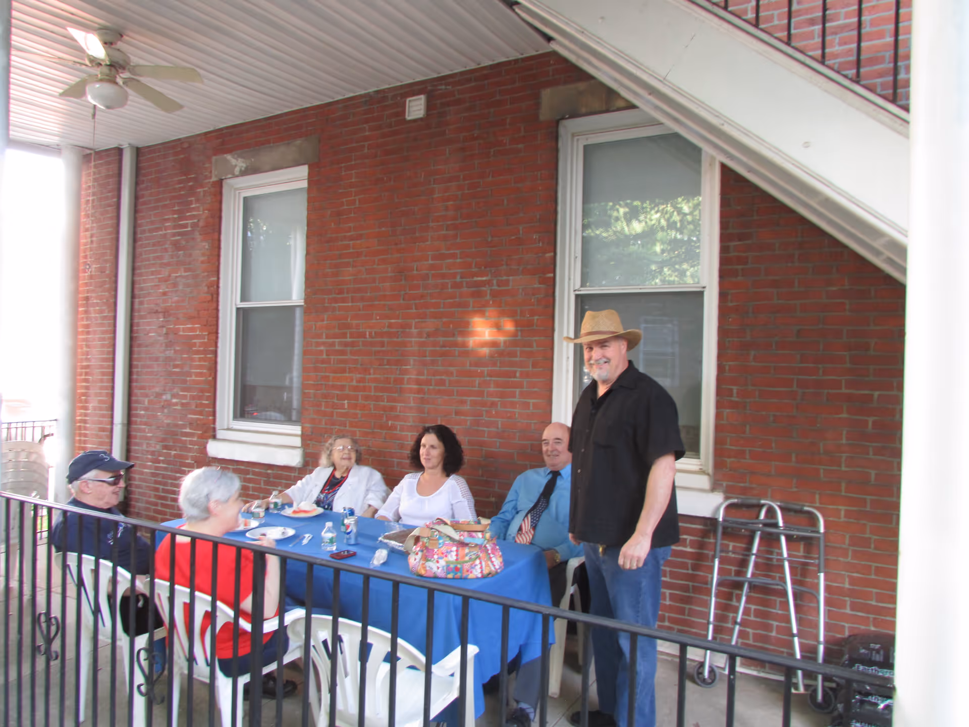 A group of five people gathered around a table with a blue tablecloth on a covered outdoor porch area with red brick walls. Four people are seated on white plastic chairs, and one man wearing a black shirt and a straw hat is standing. There is a walker leaning against the wall near the window behind them.