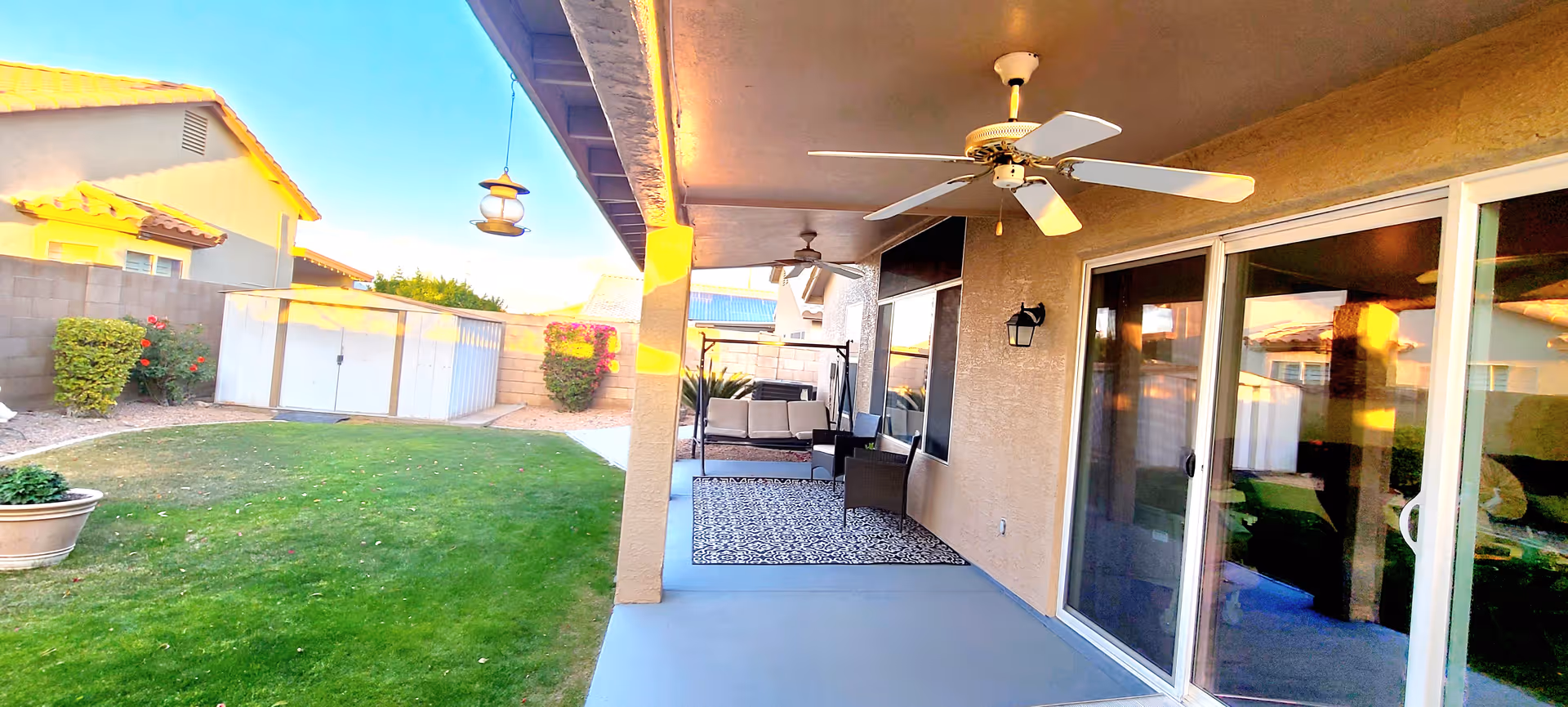Covered patio area with ceiling fans, outdoor seating including a swing and chairs on a patterned rug, adjacent to a grassy backyard with bushes, a storage shed, and neighboring houses visible in the background under a clear blue sky.