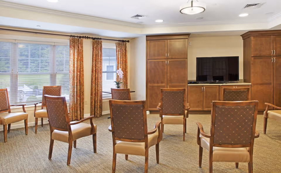 A common area with several wooden chairs arranged in a semi-circle facing a flat-screen TV mounted on a wooden cabinet. The room has large windows with patterned curtains, beige walls, and carpeted flooring.