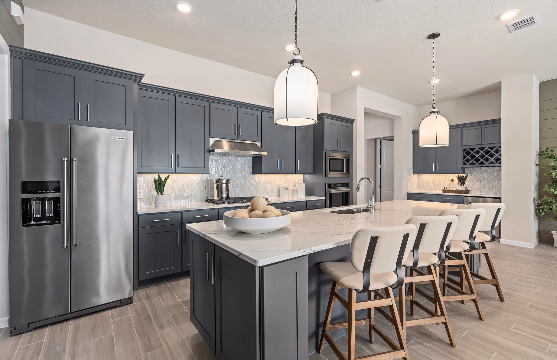 Modern kitchen with gray cabinets, a large white marble island with four beige cushioned bar stools, stainless steel refrigerator, built-in oven and microwave, two pendant lights hanging above the island, and light wood flooring.