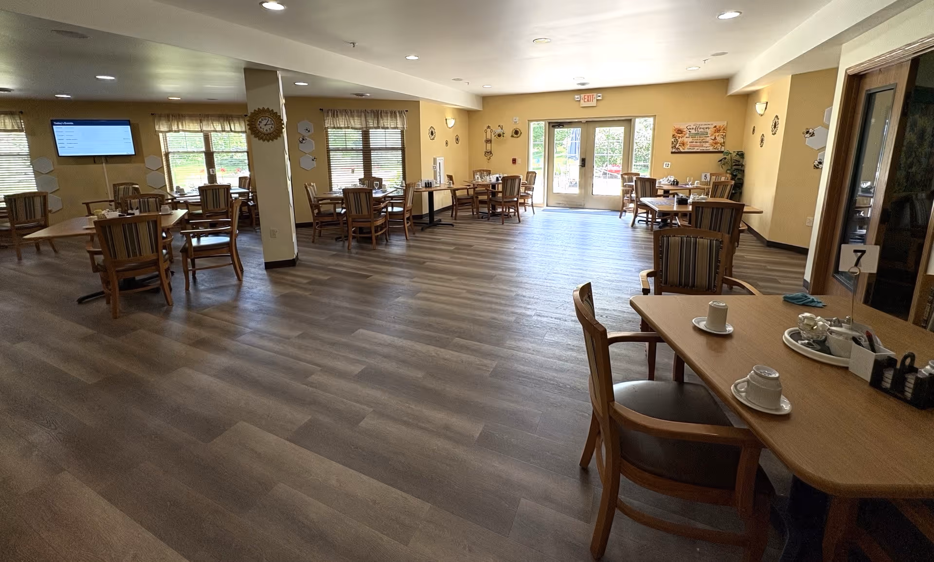 A spacious dining room with multiple wooden tables and chairs arranged neatly. The room has large windows with curtains allowing natural light to enter. There is a digital screen mounted on the wall and a clock on a pillar. The floor is covered with wood-patterned vinyl or laminate. The room has a warm, inviting atmosphere with beige walls and decorative wall hangings. A set of double glass doors leads outside.