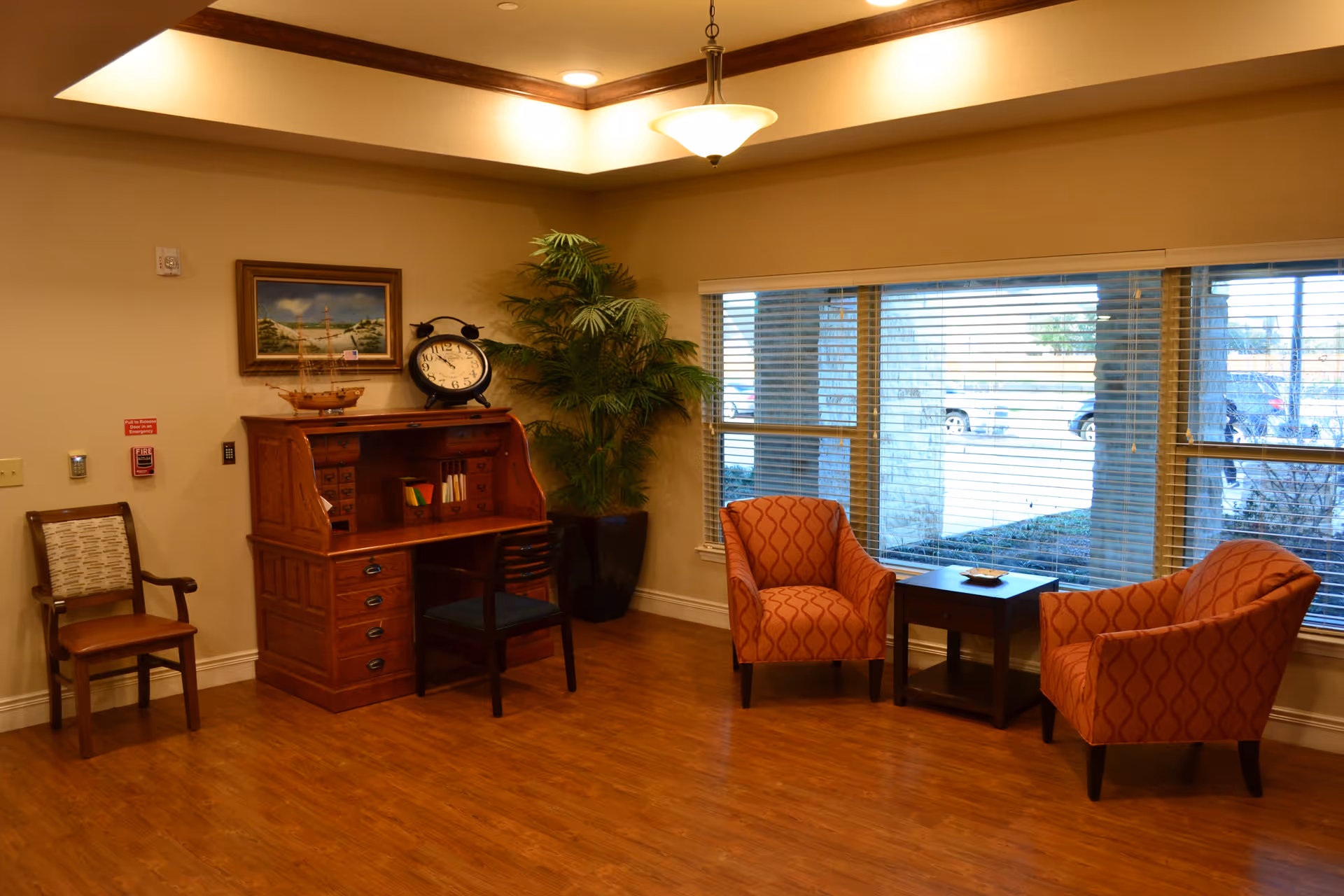 A cozy seating area with two patterned armchairs, a small table, a wooden roll-top desk, and a large window with blinds.