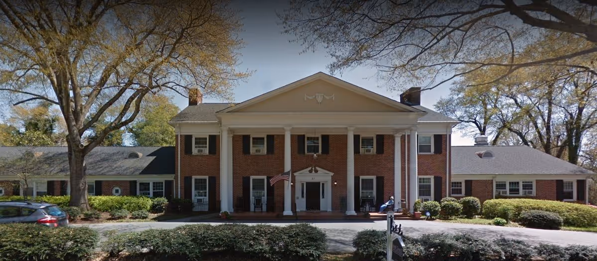 Front exterior view of a large two-story brick building with white columns and a triangular pediment above the entrance. The building is surrounded by trees and shrubs, with a driveway and parked car visible in front.