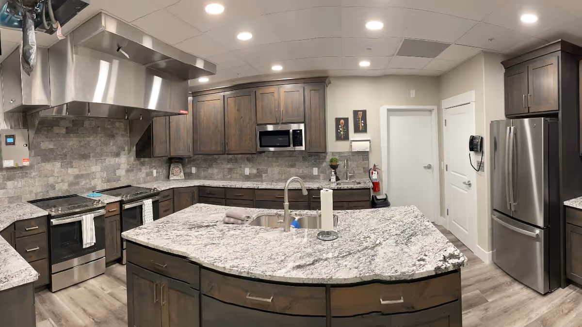 A modern kitchen with dark wooden cabinets, granite countertops, and stainless steel appliances including a refrigerator, microwave, and two ovens. There is a large kitchen island with a sink and paper towel holder. The kitchen has a tiled backsplash and recessed ceiling lights.