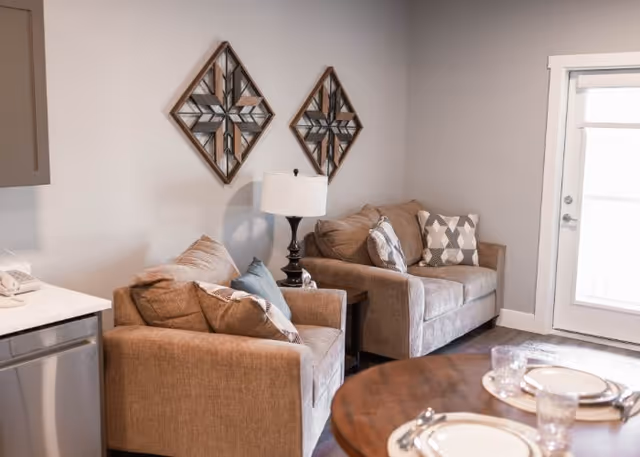 A cozy living room area with two beige sofas adorned with patterned throw pillows, a dark wooden side table with a lamp, and two decorative wall mirrors. A round wooden dining table with place settings is partially visible in the foreground, and a glass door leading outside is on the right.