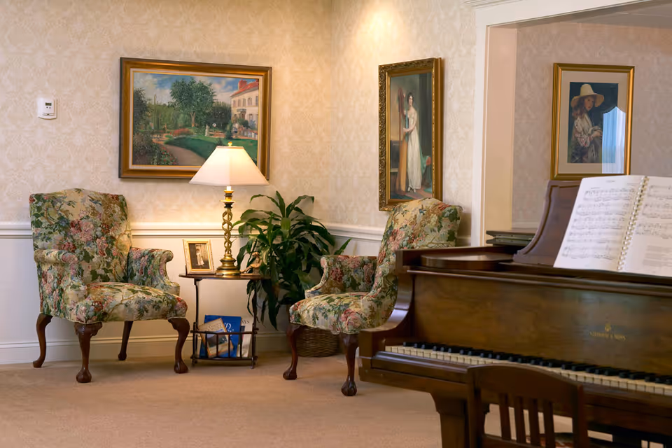 A cozy sitting area with two floral upholstered armchairs, a small side table with a lamp and framed photo, a potted plant, and three framed paintings on the wallpapered walls. A grand piano with sheet music is partially visible in the foreground.