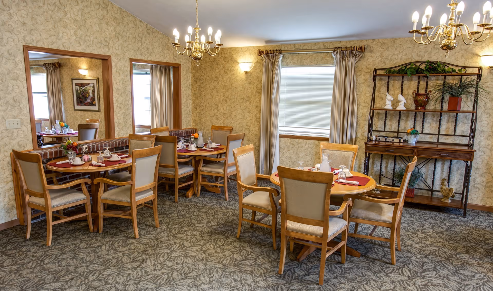 A dining room in a senior living facility with round wooden tables set with placemats, napkins, glasses, and small flower arrangements. The room has patterned wallpaper, carpeted floor, two chandeliers, a window with curtains, a large mirror on one wall, and a decorative shelving unit with plants and ornaments.