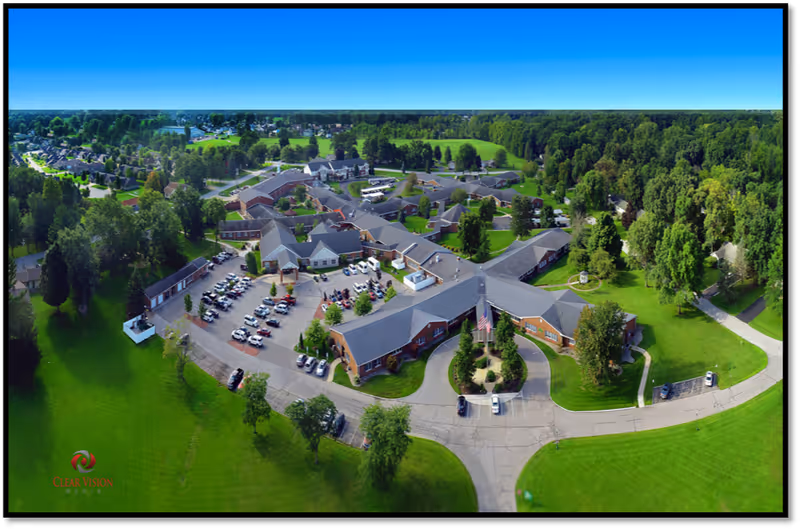 Aerial view of Grace Village Retirement Community showing multiple connected buildings with gray roofs surrounded by green lawns and trees. There is a parking lot with several cars and a circular driveway with an American flag in the center. The surrounding area includes residential houses and dense greenery under a clear blue sky.