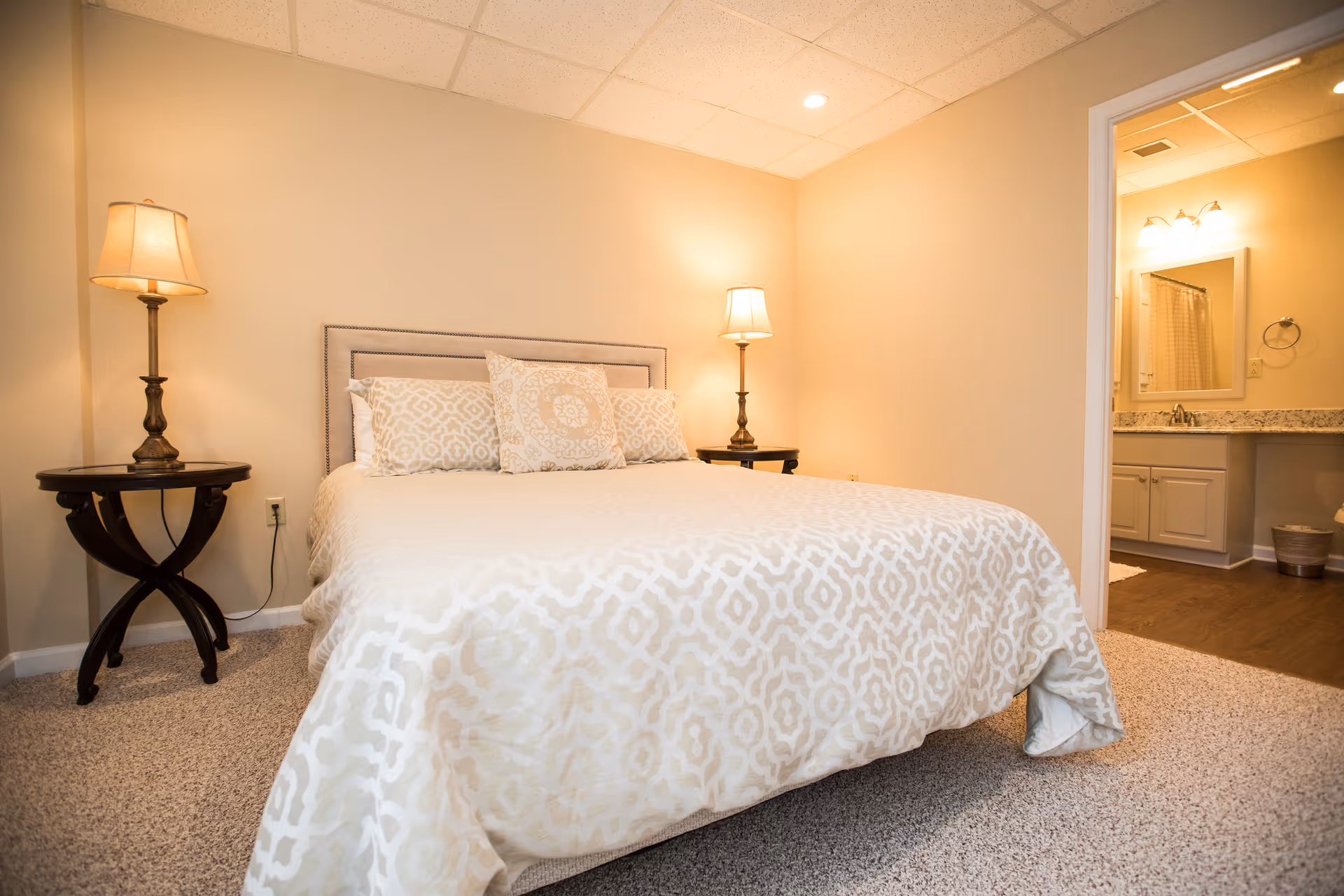 A cozy bedroom with a beige patterned bedspread on a bed, two matching pillows, and two bedside tables each with a lamp. The room has beige walls and carpeted flooring. An open doorway reveals a bathroom with a granite countertop, a mirror, and a shower curtain.